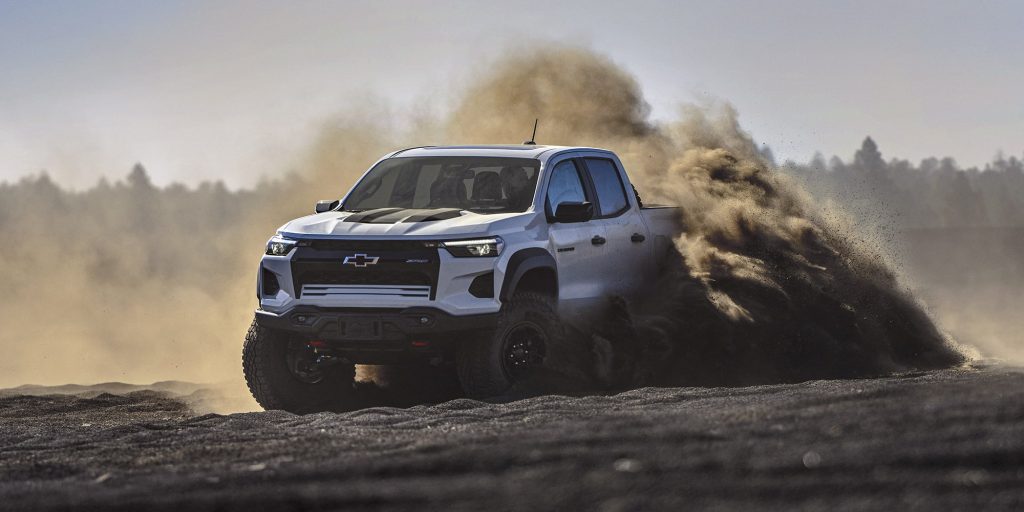 A white Chevrolet Colorado ZRX pickup truck driving off-road, kicking up a large cloud of dark brown dust against a hazy, tree-lined background.