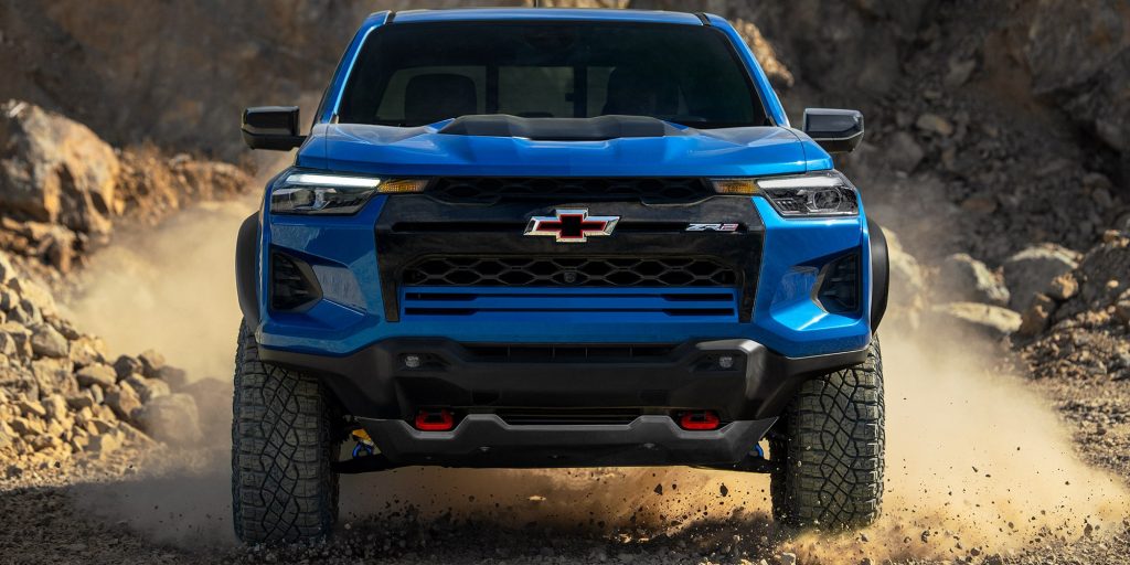 A blue Chevrolet Colorado ZR2 pickup truck driving on a dusty, rocky terrain, with dust swirling around its large off-road tires.