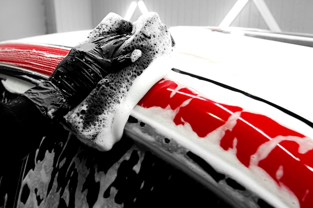 Professional car detailing process showing hand washing with a foam-covered sponge on a red vehicle, emphasizing exterior cleaning, paint protection, and auto detailing services.