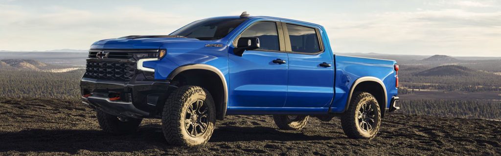 A bright blue Chevrolet Silverado ZR2 pickup truck parked on a rocky, elevated overlook with a vast forest landscape below.