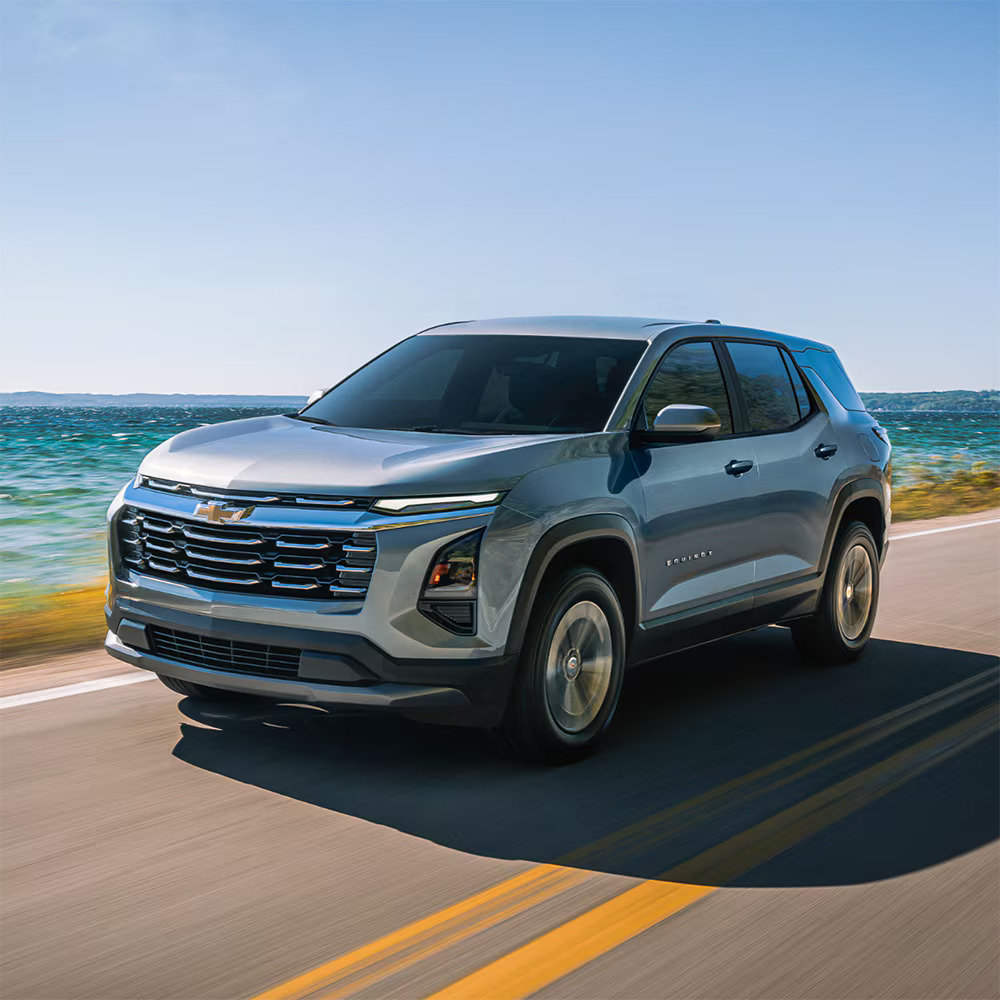 A silver Chevrolet Equinox SUV driving along a coastal highway next to a blue ocean under a clear sky.